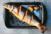 A whole salmon with roasted dill sauce and lemon halves on a metal baking tray.