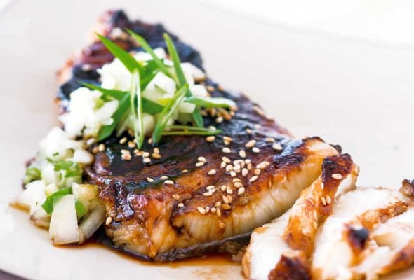 A piece of sea bass topped with soy glaze and cucumber salsa on a square shaped white plate.