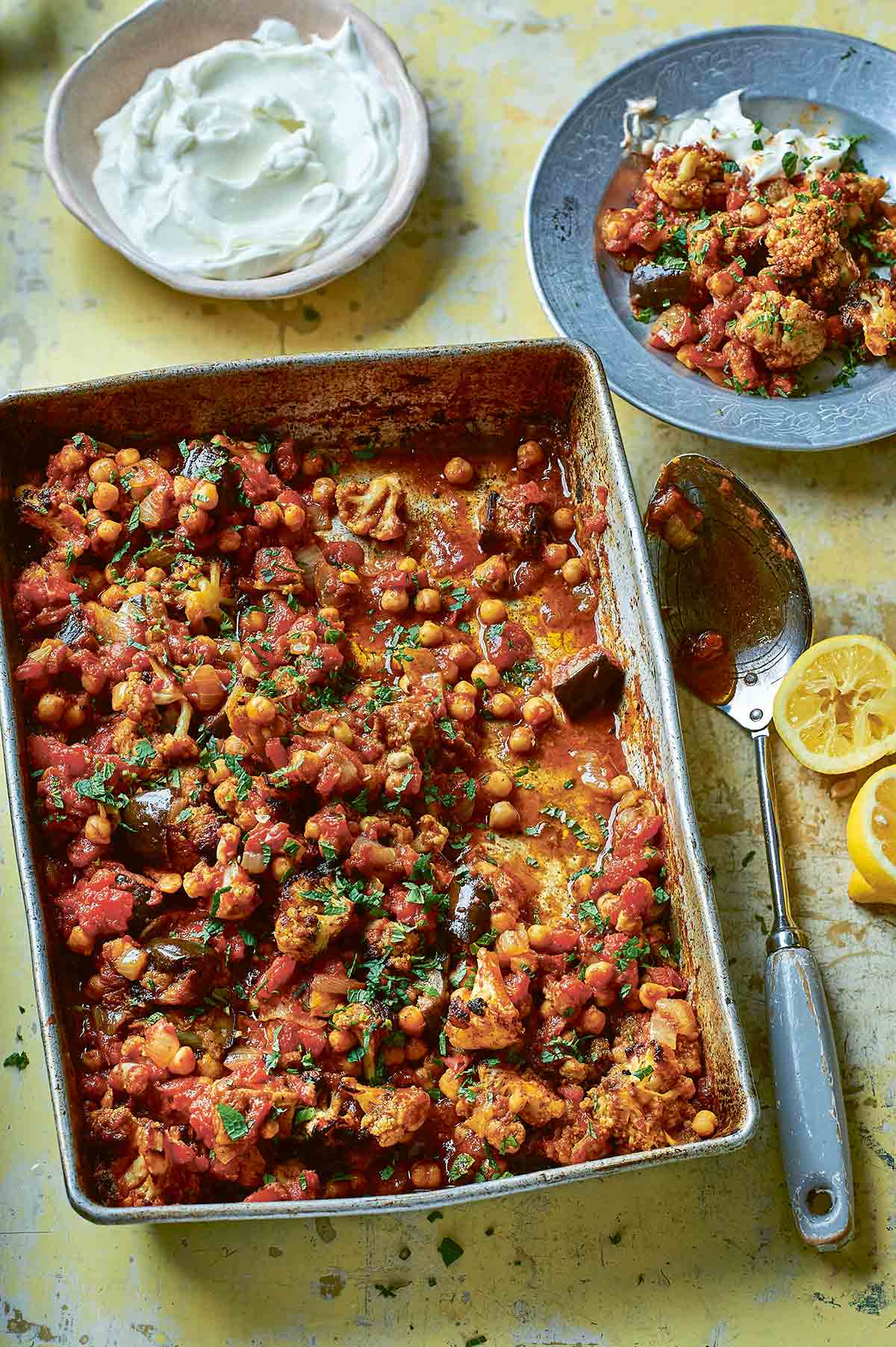 A metal baking tray filled with sheet pan chickpea curry with a spoon, two lemon halves, and a bowl of Greek yogurt beside it.
