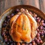 A whole simple roast chicken in a skillet, surrounded by grapes.