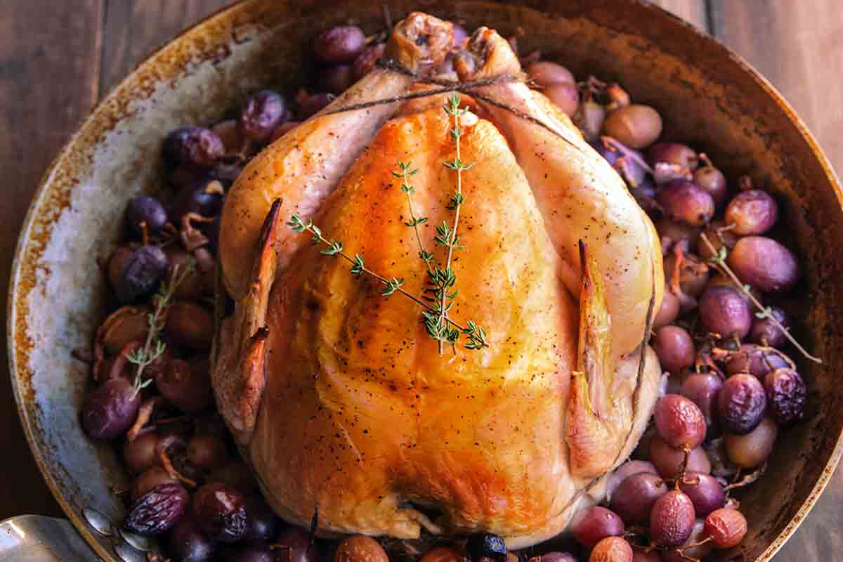 A whole simple roast chicken in a skillet, surrounded by grapes.