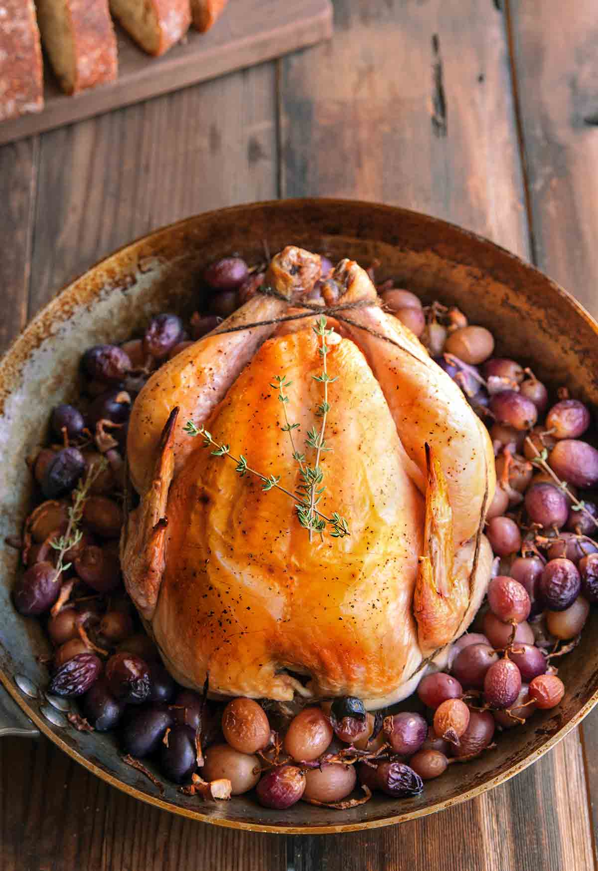 A whole simple roast chicken in a skillet, surrounded by grapes.