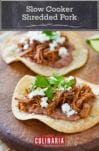 Two corn tortillas topped with slow cooker shredded pork, cheese, and cilantro on a wooden board with some lime wedges beside.