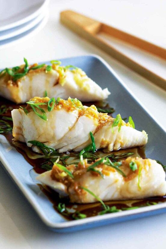 Three pieces of steamed halibut with ginger and scallions on a blue rectangular plate with chopsticks on the side.
