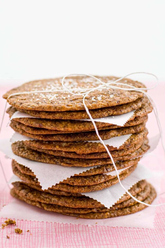 A stack of thin and crisp chocolate chip cookies with pieces of parchment in between, tied up with string and a glass bottle of milk in the background.