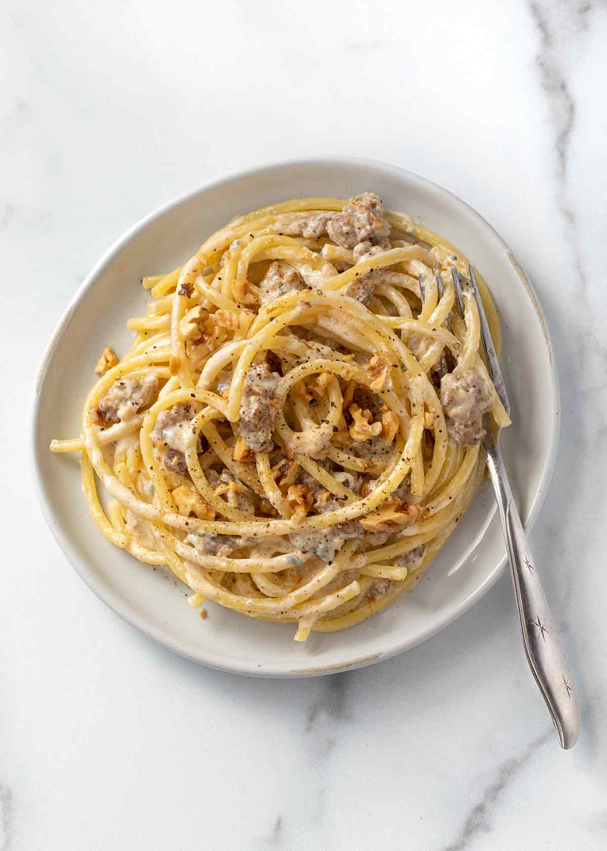 A white bowl filled with blue cheese, sausage, and walnut pasta with a fork resting on the side of the bowl.