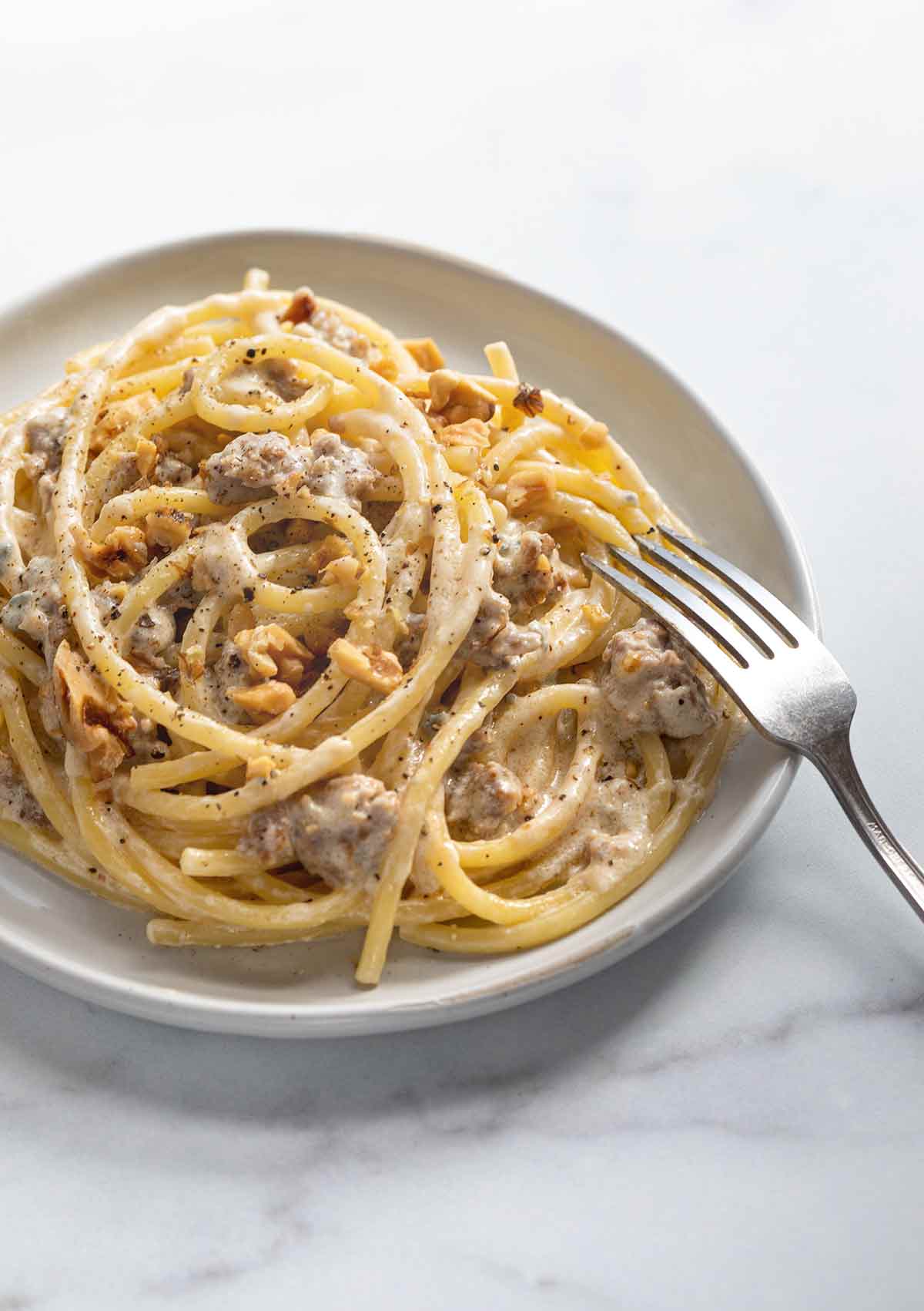 A white bowl filled with blue cheese, sausage, and walnut pasta with a fork resting on the side of the bowl.