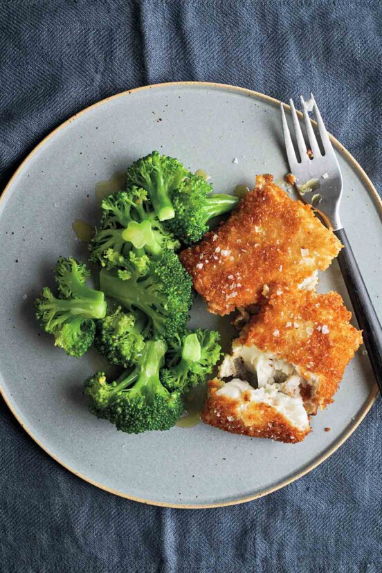 Two pieces of breaded fish fillets sprinkled with salt on a grey plate with a portion of steamed broccoli and a fork.