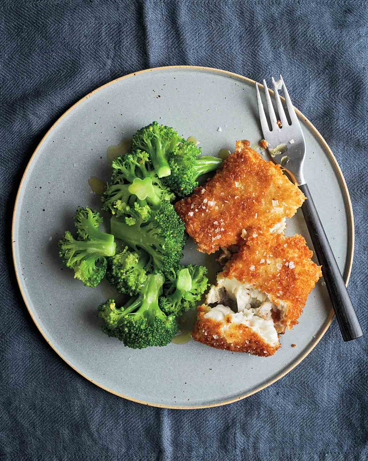 Two pieces of breaded fish fillets sprinkled with salt on a grey plate with a portion of steamed broccoli and a fork.