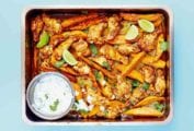 A metal tray filled with chipotle chicken wings, sweet potato wedges, and a Greek yogurt dipping sauce with lime wedges and cilantro leaves.