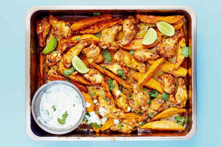 A metal tray filled with chipotle chicken wings, sweet potato wedges, and a Greek yogurt dipping sauce with lime wedges and cilantro leaves.