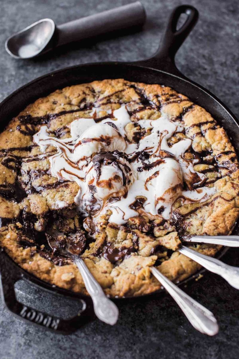 A chocolate chunk skillet cookie topped with ice cream and chocolate sauce in a cast-iron skillet with four spoons resting in the cookie.