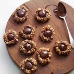 Nine chocolate thumbprint cookies and a chocolate covered spoon on a round wooden board.