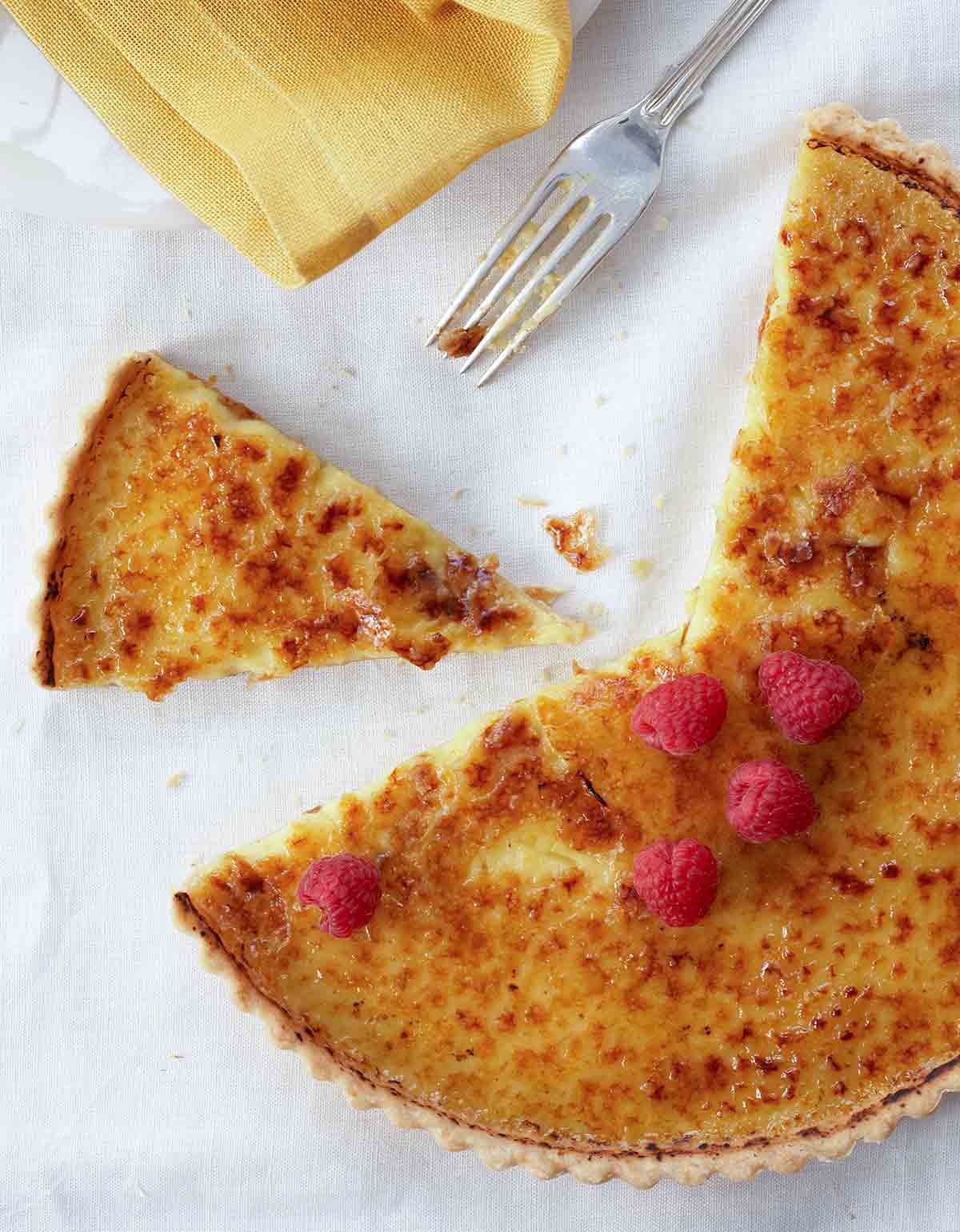 A partially sliced coconut crème brûlée tart topped with raspberries on a white linen cloth with a fork and napkin beside it.