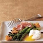 Grilled asparagus, prosciutto, fried bread, and poached egg on a white plate with a fork resting on the side.