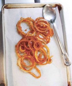A pile of homemade onion rings on a parchment-lined baking sheet with a spoon resting beside them.