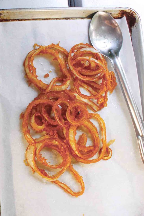 A pile of homemade onion rings on a parchment-lined baking sheet with a spoon resting beside them.