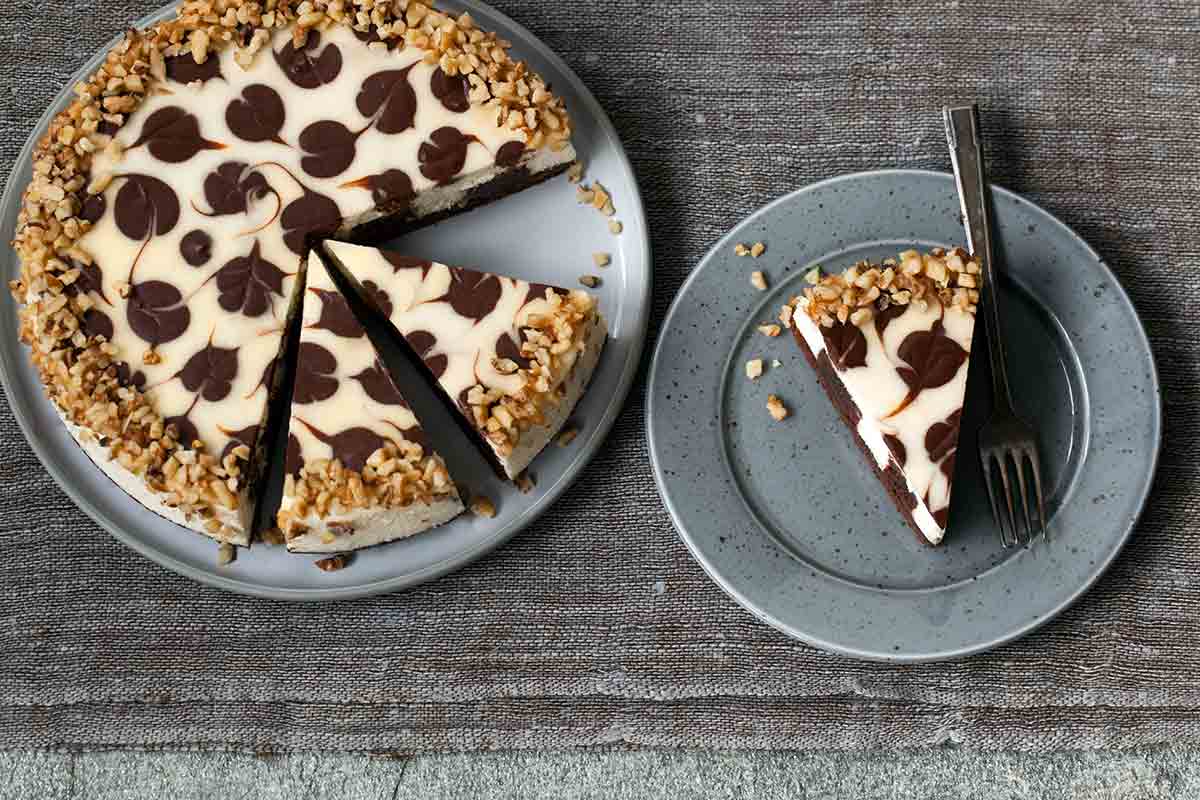 A whole Junior's brownie swirl cheesecake with three slices cut from it, and one of those slices on a grey speckled plate with a fork resting beside.