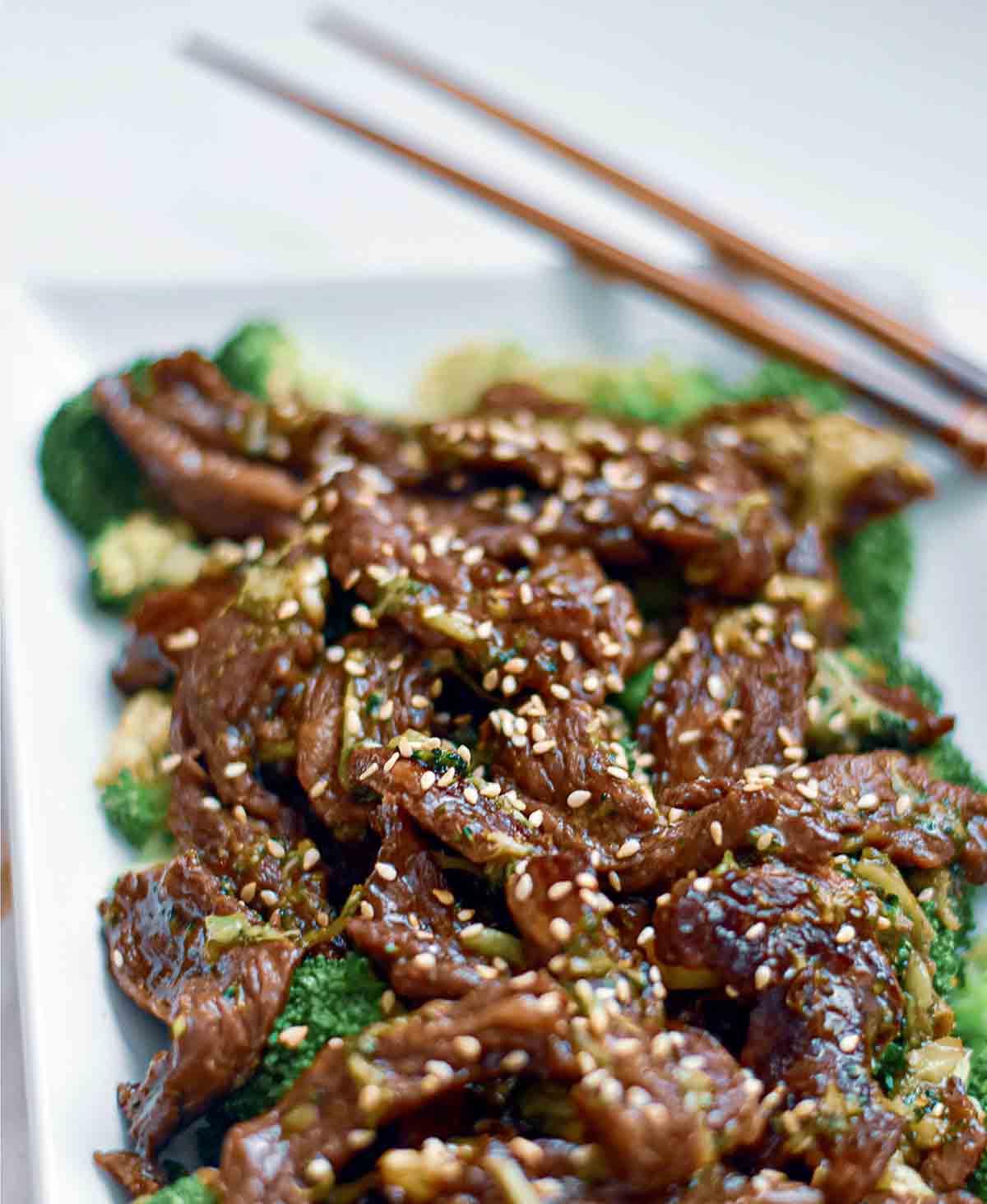 A white rectangular platter filled with keto beef and broccoli and sprinkled with sesame seeds with a pair of chopsticks resting on the edge of the platter.
