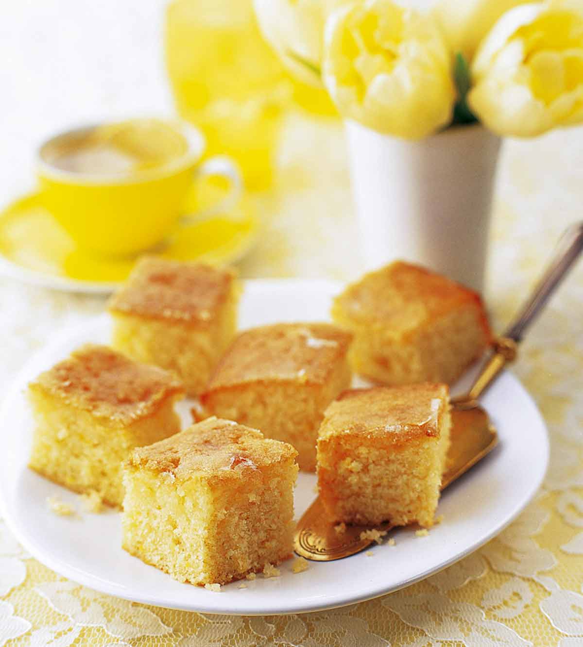 Squares of lemon drizzle cake on a white plate with a cake server resting underneath and a cup of tea and vase of flowers in the background.
