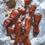 Several strips of maple bacon on a sheet of parchment, with a pastry brush and drizzles of maple syrup beside the bacon.