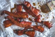 Several strips of maple bacon on a sheet of parchment, with a pastry brush and drizzles of maple syrup beside the bacon.