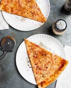 Two slices of New York style pizza on paper plates with a cheese shaker and napkins beside the pizza.
