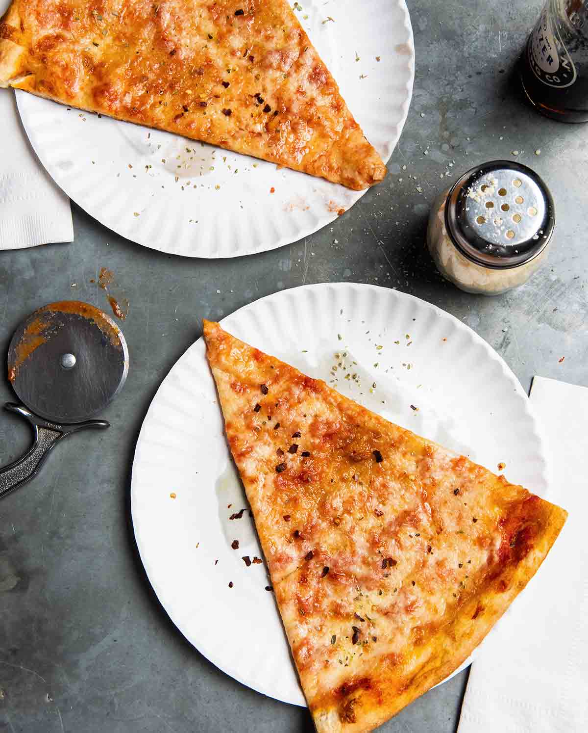 Two slices of New York style pizza on paper plates with a cheese shaker and napkins next to the pizza.