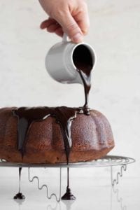 A person pouring glaze over an orange chocolate chunk cake on a round wire rack.
