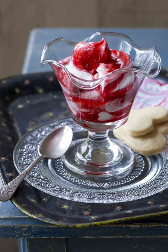 A parfait glass filled with raspberry fool on a decorative glass plate with a silver spoon and two heart-shaped cookies.