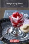 A parfait glass filled with raspberry fool on a decorative glass plate with a silver spoon and two heart-shaped cookies.