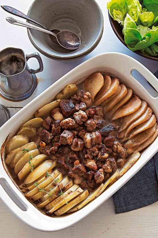 A white casserole dish of roast pork, potatoes and pears with a bowl of salad and two empty bowls with spoons beside it.