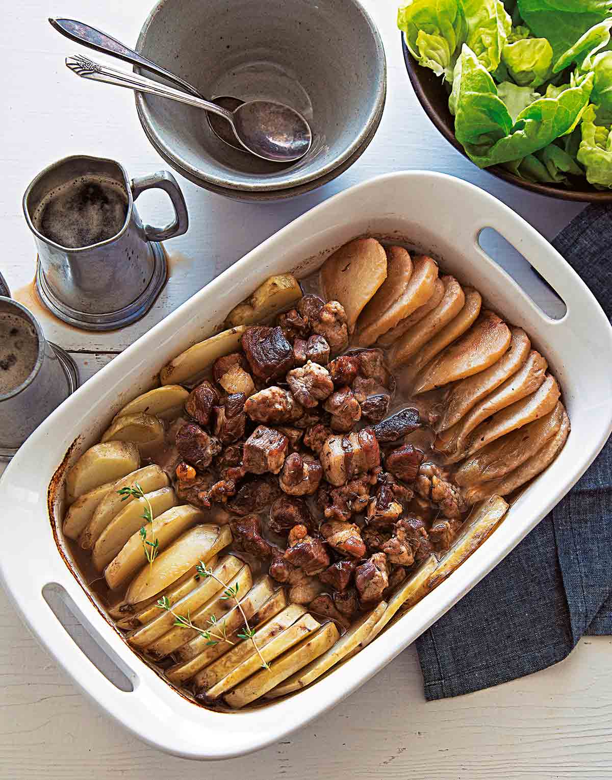 A white casserole dish of roast pork, potatoes and pears with a bowl of salad and two empty bowls with spoons beside it.