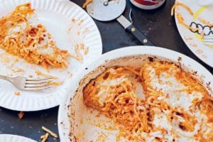 A white dish with a partially cut spaghetti pie, two paper plates with slices of spaghetti pie, a pizza cutter, napkins, and two cans of beer.