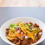 Spicy beef taco bake with ground beef, beans tortilla chips, chopped onions, and lettuce in a white bowl.