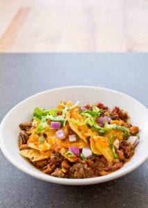 Spicy beef taco bake with ground beef, beans tortilla chips, chopped onions, and lettuce in a white bowl.