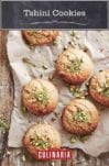 Six tahini cookies on a piece of parchment with chopped pistachios on the cookies and on the parchment.