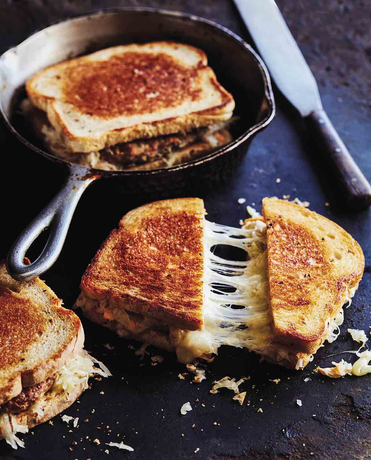 Three turkey reuben patty melts - one in a cast-iron skillet, and two on a dark surface, one sliced diagonally.