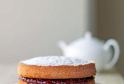 A two-tier Victoria sponge cake with raspberry jam in the middle, all dusted with confectioners' sugar on a white plate with a white teapot in the background