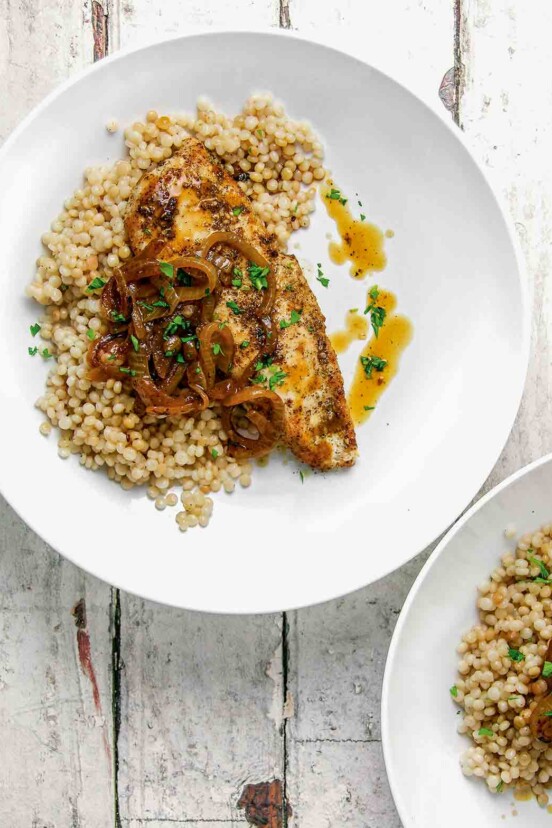 Two white plates each topped with a serving of za'atar chicken, Israeli couscous, sauteed onions, and pan juices.