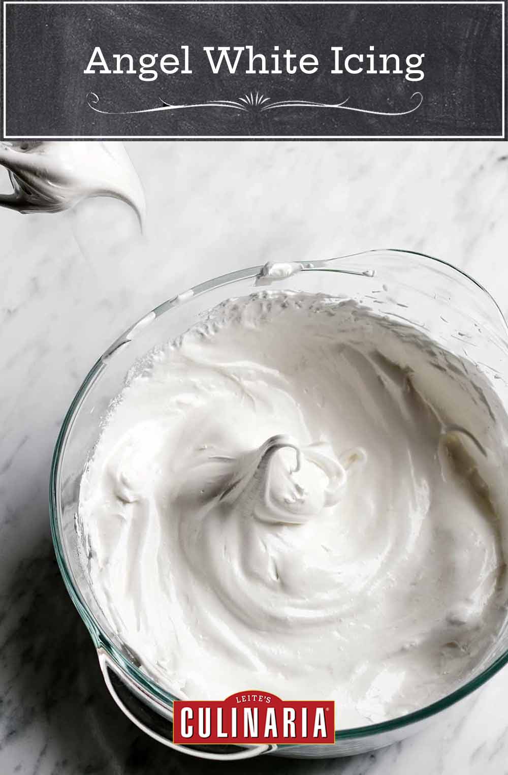 Bowl of angel white icing in firm peaks on white marble, above is a whisk