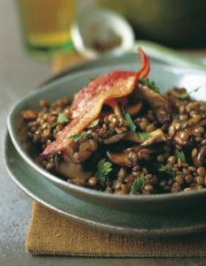 A green bowl filled with sautéed bacon, mushrooms, and lentils with a whole piece of bacon on top.