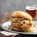 A beer battered chicken sandwich topped with slaw on a white plate on top of a paper napkin on a wooden table with a glass of beer in the background.