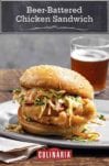 A beer battered chicken sandwich topped with slaw on a white plate on top of a paper napkin on a wooden table with a glass of beer in the background.