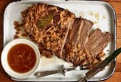 An enamel baking dish holding a sliced oven brisket topped with onions with jus on the side.