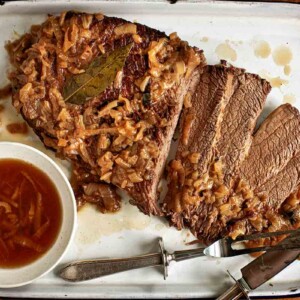 An enamel baking dish holding a sliced oven brisket topped with onions with jus on the side.