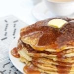 A stack of gluten-free pancakes with butter and maple syrup on a white plate on top of a crossword puzzle.