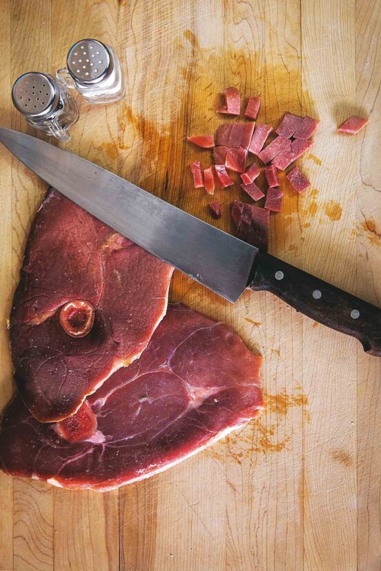 A slice of ham with red eye gravy on a cutting board with a knife and salt and pepper shaker.