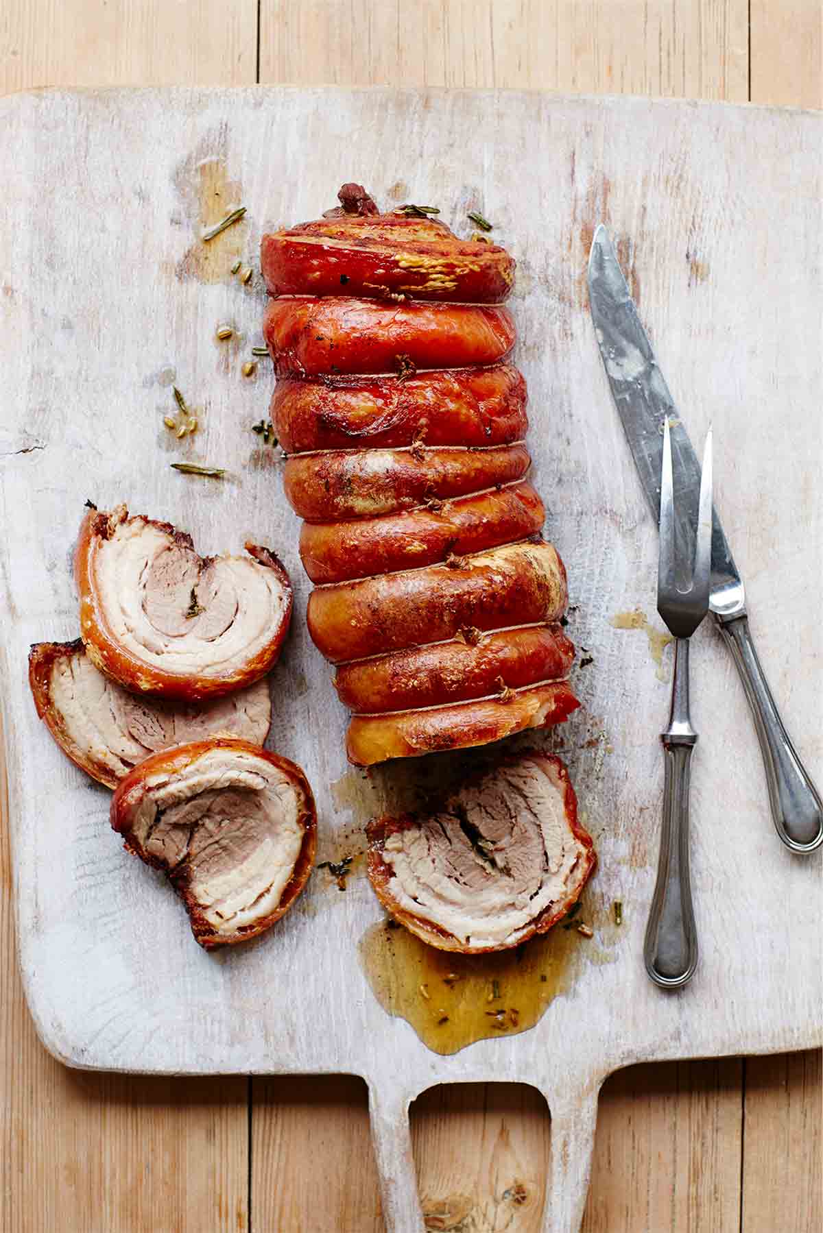 A pork belly porchetta on a cutting board with four slices carved from the end and a carving fork and knife resting beside the meat.