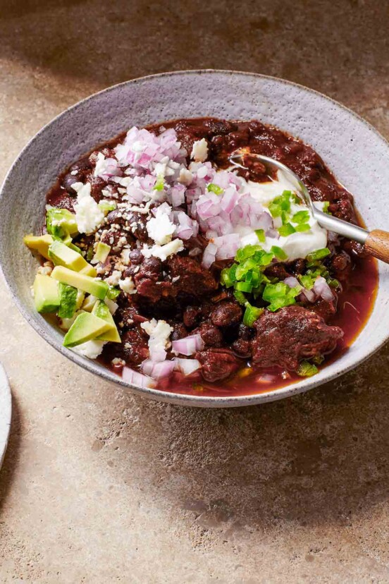 A white bowl filled with beef and black bean chili, topped with red onion, avocado, queso fresco, and diced jalapeno